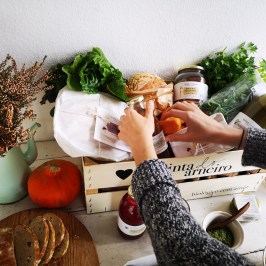 Person arranging fresh produce and jars in a wooden crate on a wooden table.