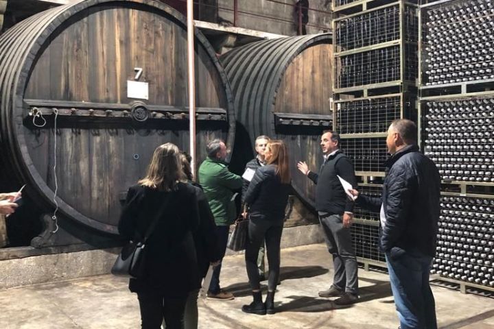 People touring a winery with large wooden barrels and stacked wine bottles.