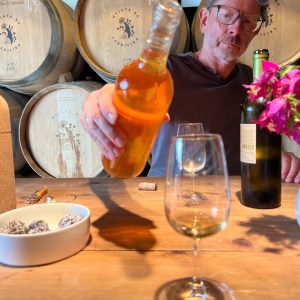 Man pours amber liquid into glass, surrounded by wine barrels and flowers.