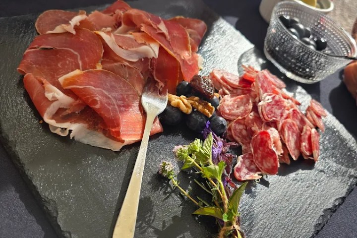 Charcuterie board with cured meats and herbs on a slate, with olives and wine in the background.