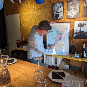Man examining a framed map on a shelf in a cozy room with wine glasses on a table.