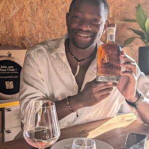 Smiling person holding a bottle at a table with wine glasses.