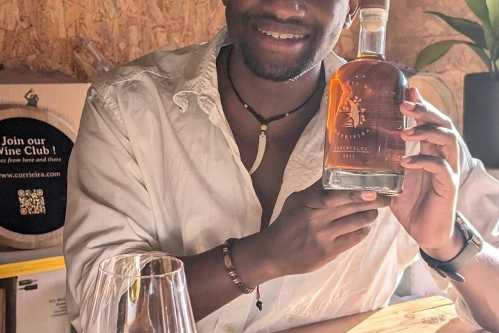 Smiling person holding a bottle at a table with wine glasses.