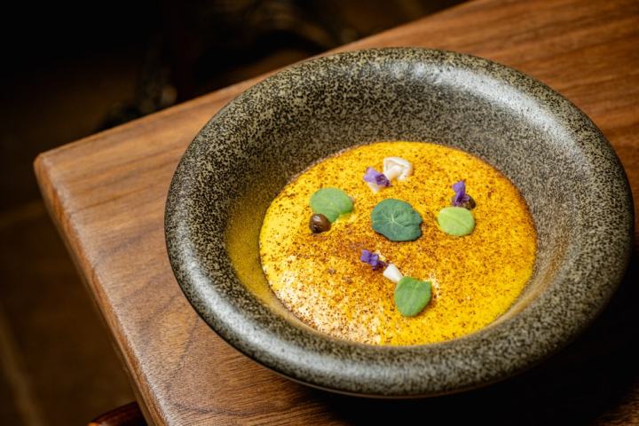 Elegant dish with yellow sauce, herbs, and edible flowers in a stone bowl on wooden table.