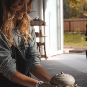 Smiling woman potter shaping clay on a wheel in a sunlit room with open door.