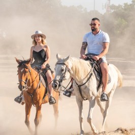Two people riding horses on a dusty trail, one wearing a hat.