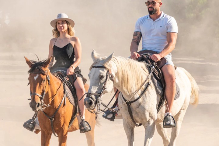 Two people riding horses on a dusty trail, one wearing a hat.