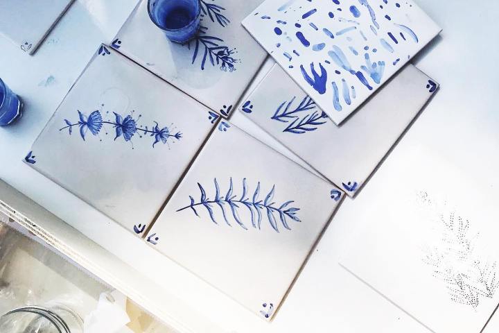 White tiles with blue plant and abstract designs on a table, surrounded by painting supplies.