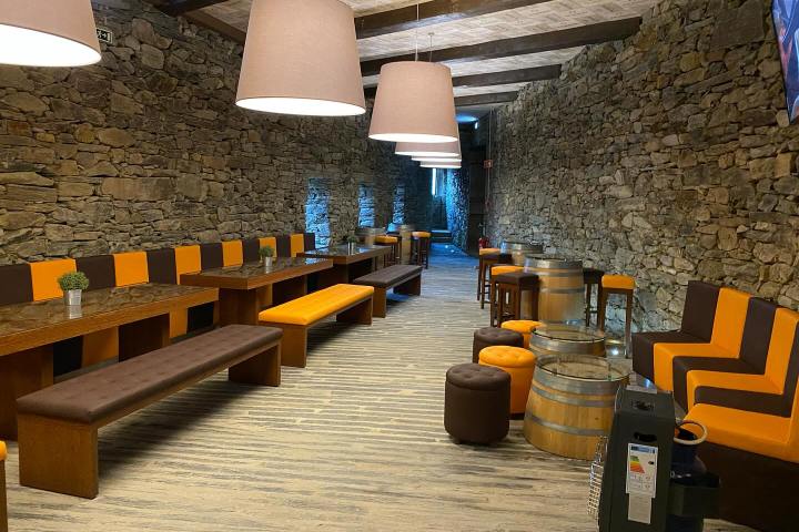 Cozy room with stone walls, wooden tables, barrel tables, orange-brown seating, and hanging lamps.