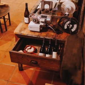 Workshop table with tools, wine bottles in a drawer, and cardboard boxes on a tiled floor.