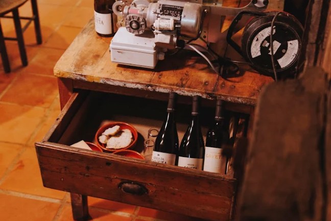 Workshop table with tools, wine bottles in a drawer, and cardboard boxes on a tiled floor.
