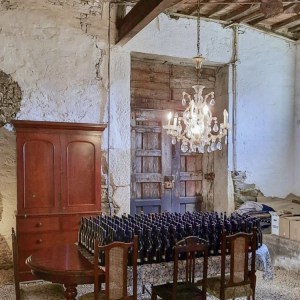 Rustic room with wooden table, chairs, cabinet, chandelier, and wine bottles in old stone building.