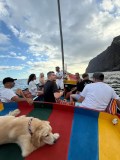 People on a colorful boat with a dog, near rocky cliffs under a partly cloudy sky.