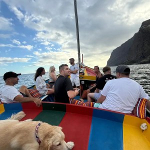 People on a colorful boat with a dog, near rocky cliffs under a partly cloudy sky.