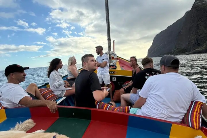 People on a colorful boat with a dog, near rocky cliffs under a partly cloudy sky.