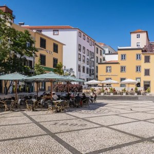 Sunny plaza with outdoor cafe tables and colorful buildings.