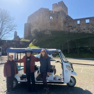 Three people posing by a tuk-tuk in front of a historic stone castle on a sunny day.