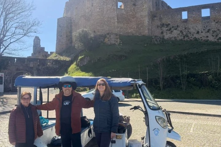 Three people posing by a tuk-tuk in front of a historic stone castle on a sunny day.