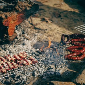 Meat skewers and sausages cooking on outdoor grills over a campfire.