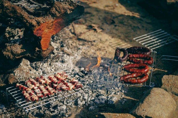 Meat skewers and sausages cooking on outdoor grills over a campfire.