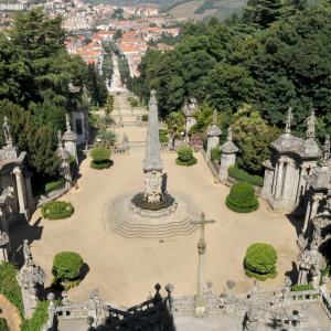 Ornate stone garden with fountain, statues, and trees, overlooking a city.