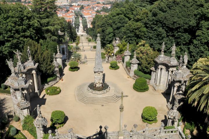 Ornate stone garden with fountain, statues, and trees, overlooking a city.