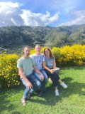 Three people sitting on a bench in front of yellow flowers and mountains.