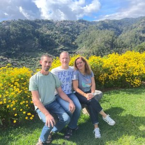 Three people sitting on a bench in front of yellow flowers and mountains.