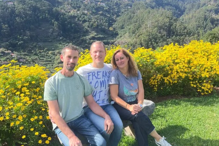 Three people sitting on a bench in front of yellow flowers and mountains.