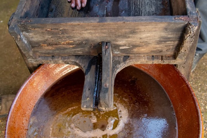 Hands pressing sack in wooden box over brown liquid in bowl.