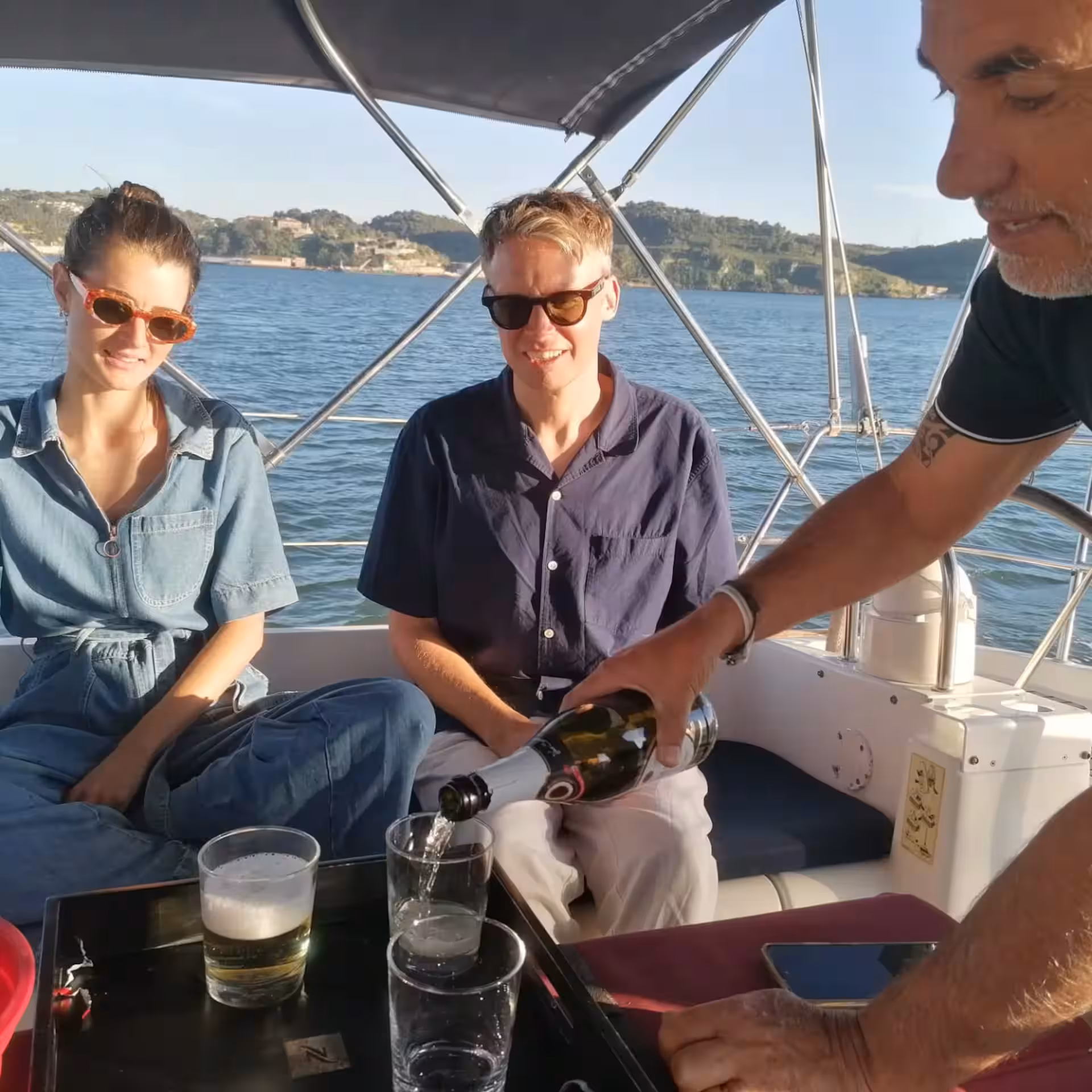 Three people on a sailboat, one pouring champagne, with a scenic water view.