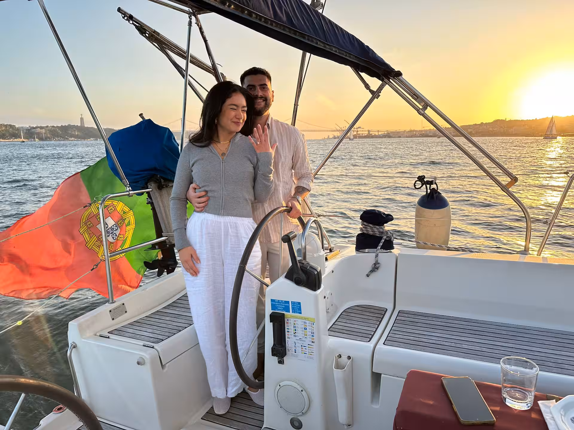 Couple on a sailboat at sunset, with a flag and water in the background.