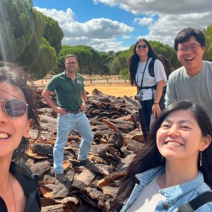 Group of people smiling outdoors near a pile of logs with trees in the background.