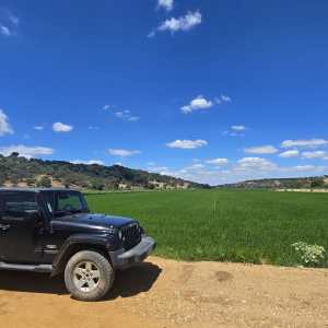 Black SUV parked on dirt road by green field under blue sky with clouds.